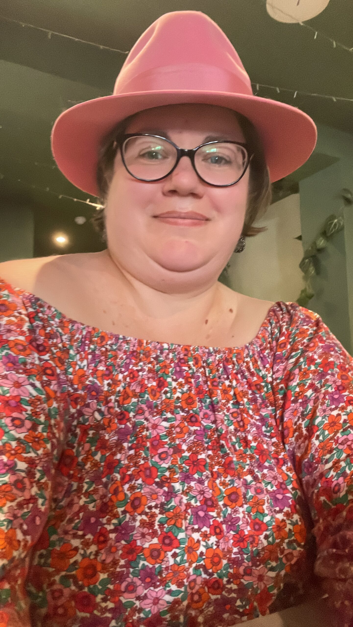 Jenn, a 40 something white woman wear a floral off the shoulder dress, pink fedora hat and black rimmed glasses